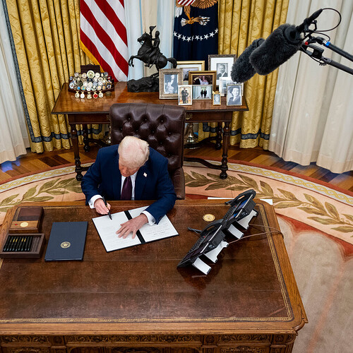President Trump sitting at his desk in the Oval Office and signing executive orders while a staffer stands nearby with more. Two boom mics are over his head.500x500